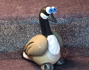 May include: A hand-crafted figurine of a goose with large blue eyes, a black neck, and a white chest. The goose is made of clay and has a realistic appearance.