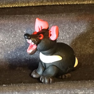 May include: A small black and white figurine of a Tasmanian devil with red ears and eyes. The devil is sitting on its haunches with its mouth open in a growl.