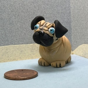 May include: A small, hand-crafted pug figurine made of clay. The figurine is beige with black ears and nose, and has blue eyes. The figurine is sitting on a light blue surface.
