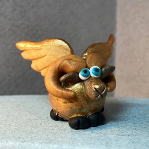 May include: A small, golden ram figurine with wings and blue eyes. The ram is made of clay and has a whimsical, cartoonish style.