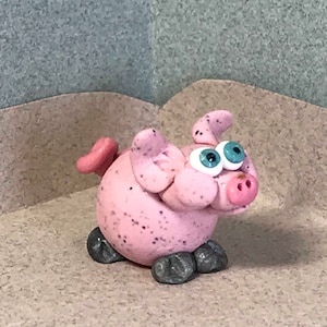 May include: A pink pig figurine with blue eyes and a speckled texture. The pig has a curled tail and gray feet.