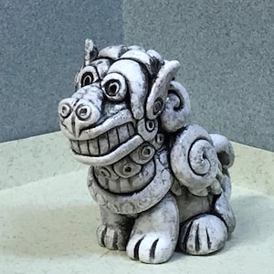 May include: A white ceramic sculpture of a mythical guardian lion, known as a Komainu, with intricate details and a fierce expression.