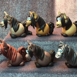 May include: Six miniature clay horses with black, brown, and gray paint. Each horse has a different color mane and tail, and all have blue eyes.