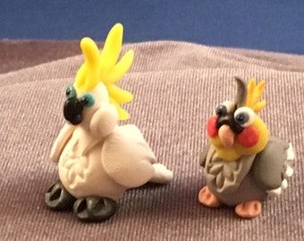 May include: Two small, hand-crafted bird figurines. The first is a white cockatoo with a yellow crest. The second is a gray cockatiel with a yellow head and red cheeks.
