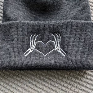 May include: Black beanie with a white embroidered design of two skeleton hands making a heart shape.
