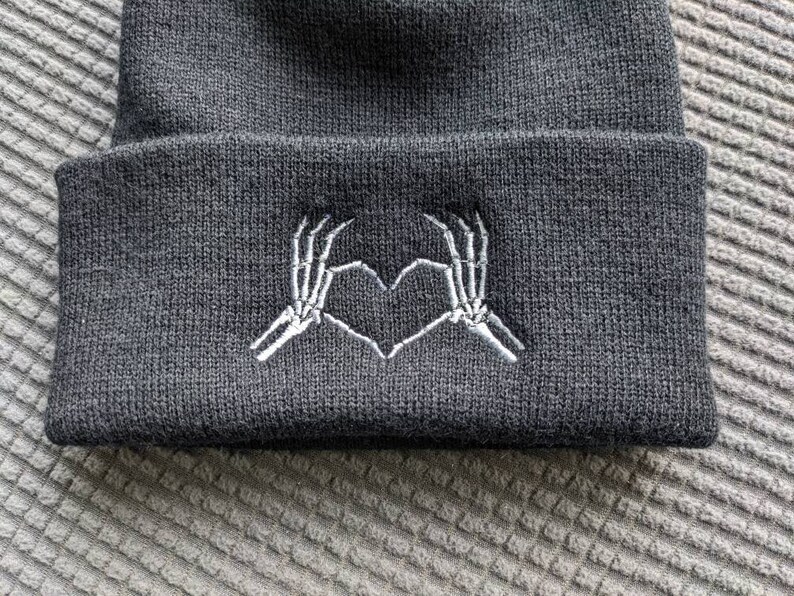 May include: Black beanie with a white embroidered design of two skeleton hands making a heart shape.