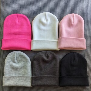May include: Six beanies in different colors: pink, white, light pink, gray, dark gray, and black. The beanies are arranged in two rows of three.