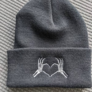 May include: Black beanie with a white embroidered design of two skeleton hands making a heart shape.