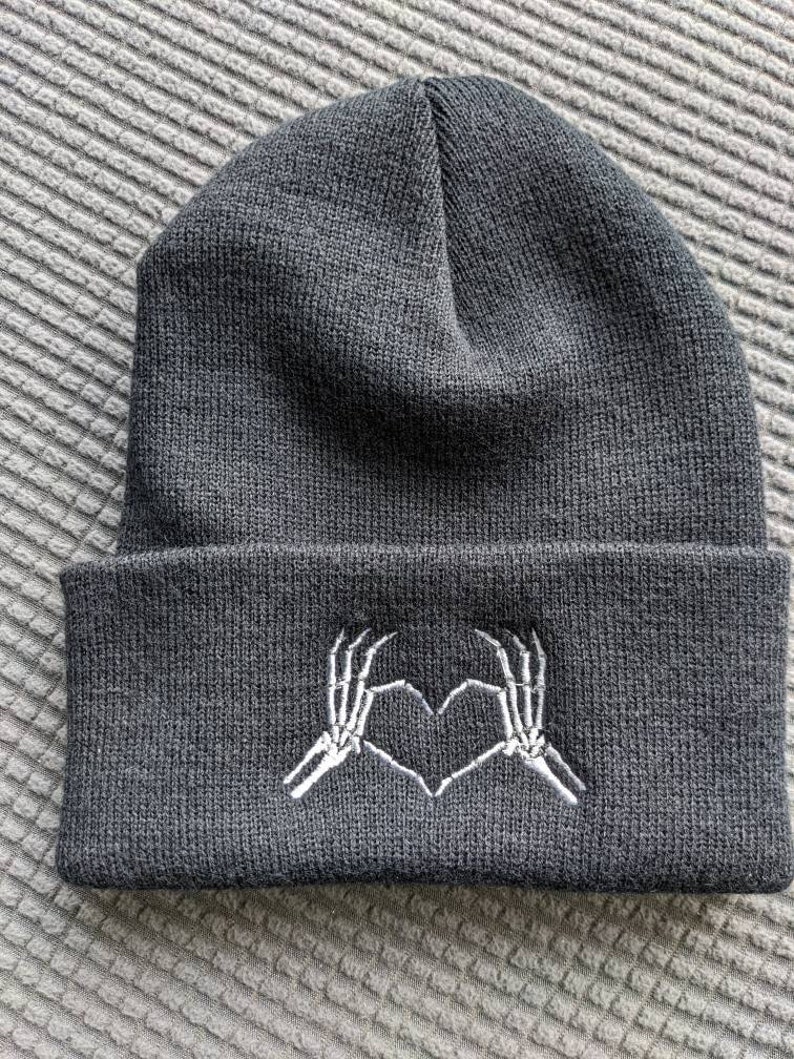 May include: Black beanie with a white embroidered design of two skeleton hands making a heart shape.