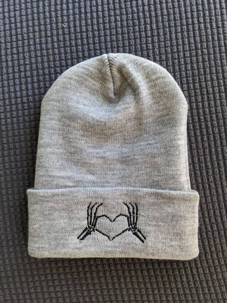 May include: Gray beanie with a black embroidered design of two skeleton hands making a heart shape.