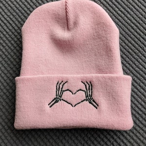 May include: A pink beanie with a black embroidered design of two skeleton hands making a heart shape.