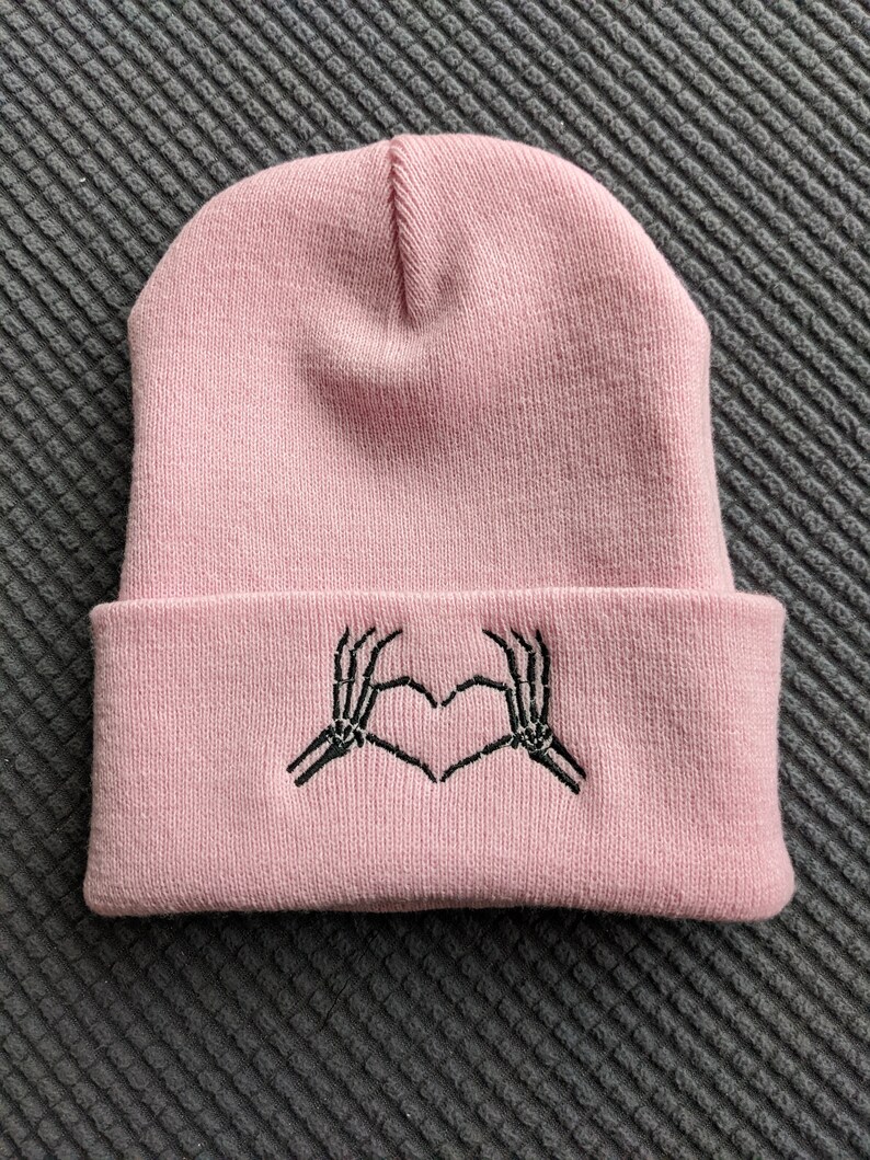 May include: A pink beanie with a black embroidered design of two skeleton hands making a heart shape.