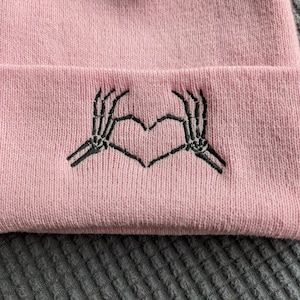 May include: Pink knit beanie with a black embroidered design of two skeleton hands making a heart shape.