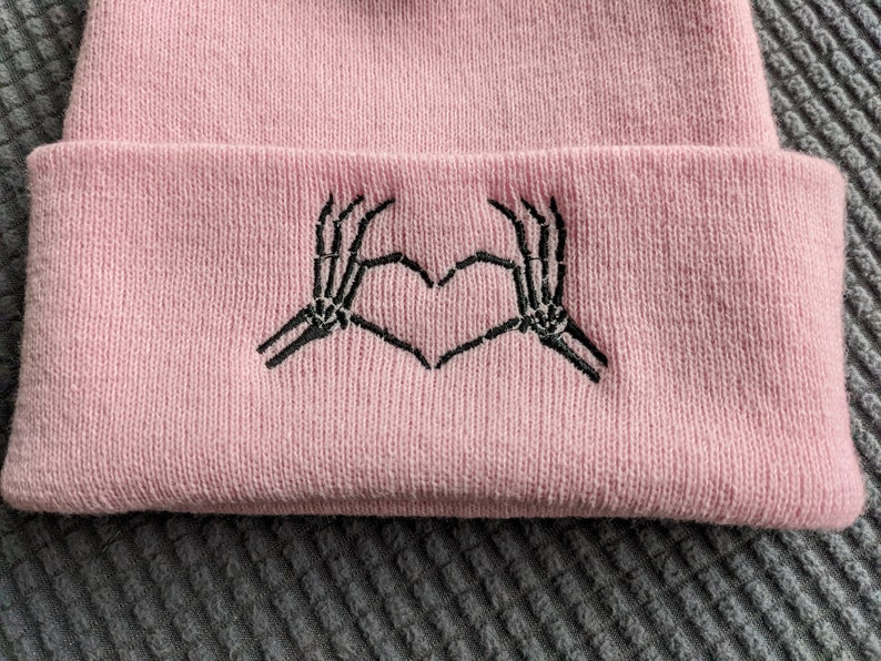 May include: Pink knit beanie with a black embroidered design of two skeleton hands making a heart shape.