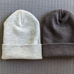 May include: Two knit beanies, one gray and one dark gray, are laid out side by side on a textured gray surface.
