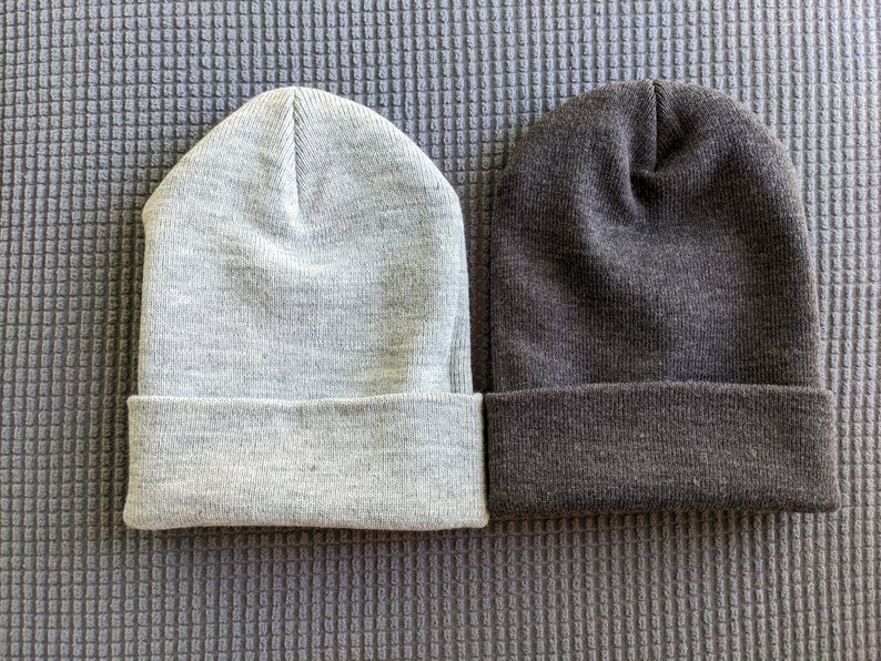 May include: Two knit beanies, one gray and one dark gray, are laid out side by side on a textured gray surface.