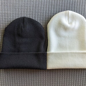 May include: Two knit beanies, one black and one white, are laid out side by side on a gray textured surface.