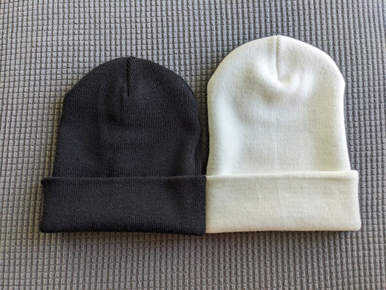 May include: Two knit beanies, one black and one white, are laid out side by side on a gray textured surface.
