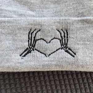 May include: Gray beanie with a black embroidered design of two skeleton hands forming a heart shape.