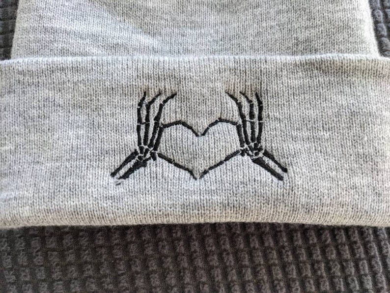 May include: Gray beanie with a black embroidered design of two skeleton hands forming a heart shape.