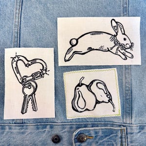 Könnte beinhalten: Hellblaue Jeansjacke mit drei Stoffaufnähern. Die Aufnäher zeigen einen herzförmigen Karabiner mit Schlüsseln, einen laufenden Hasen und zwei Birnen. Alle Designs sind schwarze Strichzeichnungen auf weißem Stoff.