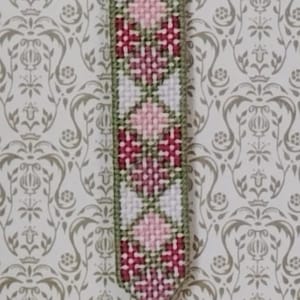 May include: Handmade bookmark with a woven design featuring pink and burgundy diamond patterns on a white background, framed by green edges. A pink tassel hangs from the bottom, and a decorative metal top.
