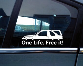 May include: A white vinyl decal applied to a car window featuring a line-art silhouette of a two-door SUV. Below the vehicle graphic, the text reads One Life. Free it! in a bold, sans-serif white font, creating a high-contrast design.
