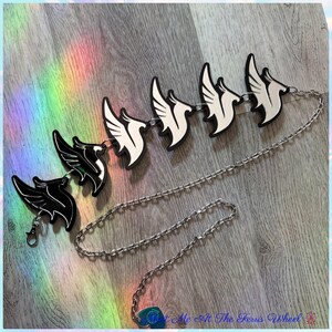 May include: A silver-toned metal chain necklace featuring six repeating wing-shaped charms. The charms alternate between solid black and solid white colors. The chain includes a lobster claw clasp and a small blue heart-shaped charm at the end.
