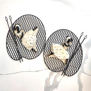 May include: Two wall art pieces featuring black metal oval frames with a lattice design. Each frame holds a figurine of a woman in a cream-colored dress with gold accents. The figurines are posed in a dancing position, with black spears extending from the frames.