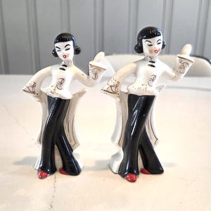 May include: Two vintage porcelain figurines of women in black pants, white tops, and red shoes. The figurines have black hair and are posed with one arm raised. The tops have gold accents. The figurines are standing on a white base.