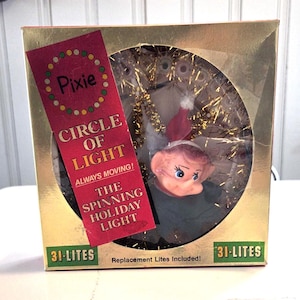 May include: A vintage Christmas decoration in a gold box. The box contains a plastic elf with a red hat and green clothing, surrounded by gold tinsel. The box has a red label that reads "Pixie Circle of Light".