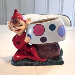 May include: A vintage ceramic planter featuring a whimsical elf figure in a red outfit, kneeling and holding a mushroom-shaped container. The mushroom is white with colorful polka dots. The base is dark green.