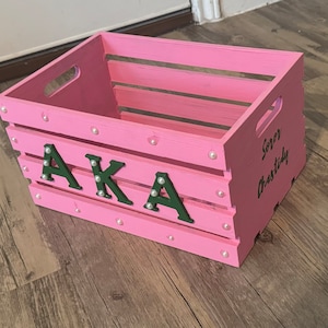 May include: A pink wooden crate with a handle on the side. The crate is decorated with white pearl-like embellishments and green letters spelling "AKA" and the text "Sorry Chastidy".