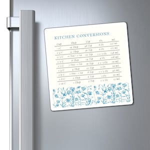 Puede incluir: Una tabla de conversión de cocina magnética blanca con detalles florales azules. La tabla enumera las medidas de cocina comunes en tazas, cucharadas, cucharaditas, onzas y mililitros.