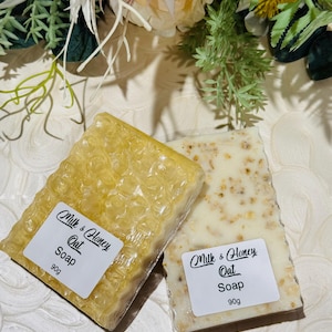 May include: Two rectangular bars of soap, one golden yellow and the other off-white, are displayed. Both soaps have labels that read "Milk & Honey Oat Soap" and "90g". The soaps are surrounded by floral arrangements with green and cream-coloured flowers.