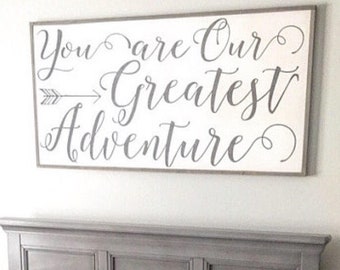 Puede incluir: Una cuna gris con una manta blanca y gris en su interior. La cuna está en una habitación de bebé con un letrero encima que dice "You are our greatest adventure".