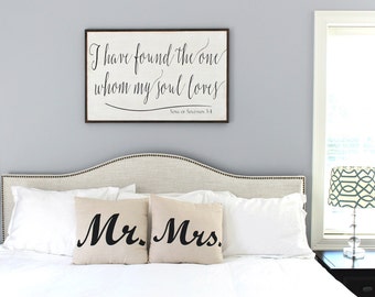 May include: A bedroom scene featuring a framed sign with the text "I have found the one whom my soul loves" and two pillows that read "Mr. Mrs." The sign is above a bed with white pillows and a neutral-toned headboard.
