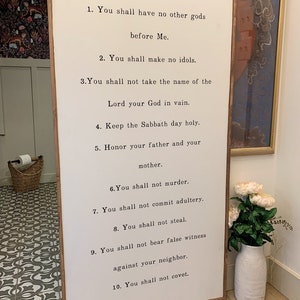 May include: White wooden sign with black text listing the Ten Commandments. The sign is hanging on a wall with a white vase with pink flowers in front of it.
