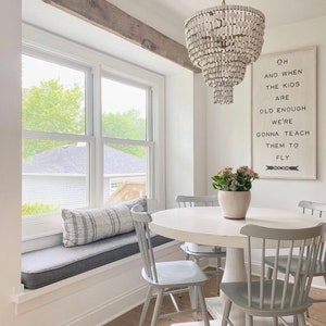 May include: A white dining table with four gray chairs sits in a room with a window seat and a wooden beam above the window. A white chandelier hangs above the table. A framed print with the text "Oh and when the kids are old enough we're gonna teach them to fly" hangs on the wall.