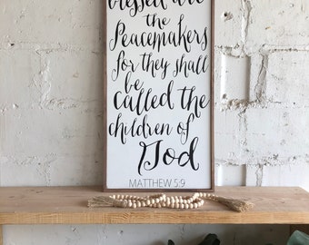 May include: A wooden sign with a white background and black lettering. The sign reads "Blessed are the Peacemakers for they shall be called the children of God" and "Matthew 5:9".