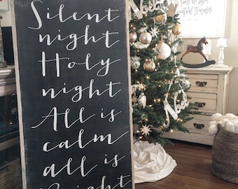 May include: A black chalkboard sign with white calligraphy that reads "Silent night Holy night All is calm all is Bright". The sign is leaning against a Christmas tree decorated with gold and silver ornaments.