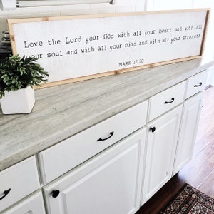 May include: A framed wood sign with a white background and black text that reads "Love the Lord your God with all your heart and with all your soul and with all your mind and with all your strength MARK 12:30". The sign is hanging on a white kitchen cabinet.