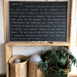 May include: A framed black sign with white text displaying Psalm 112, above a wooden shelf. The shelf holds two woven baskets with paper lanterns and a basket of green plants.