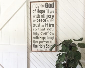 May include: A wooden sign with a white background and black text. The sign reads "may the God of Hope fill you with all joy & peace as you trust in Him so that you may overflow with Hope through the power of the Holy Spirit Romans 15:13".