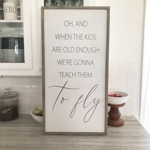 May include: A white wooden sign with a gray frame, featuring the text "OH, AND WHEN THE KIDS ARE OLD ENOUGH WE'RE GONNA TEACH THEM to fly".