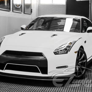 May include: A white Nissan GT-R sports car featuring a black front grille, sharp angular headlights, and dark multi-spoke alloy wheels. The vehicle has a low-profile aerodynamic design with hood vents and a sleek, modern exterior finish.
