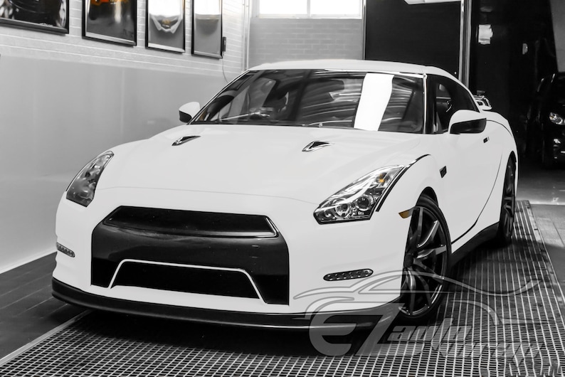 May include: A white Nissan GT-R sports car featuring a black front grille, sharp angular headlights, and dark multi-spoke alloy wheels. The vehicle has a low-profile aerodynamic design with hood vents and a sleek, modern exterior finish.