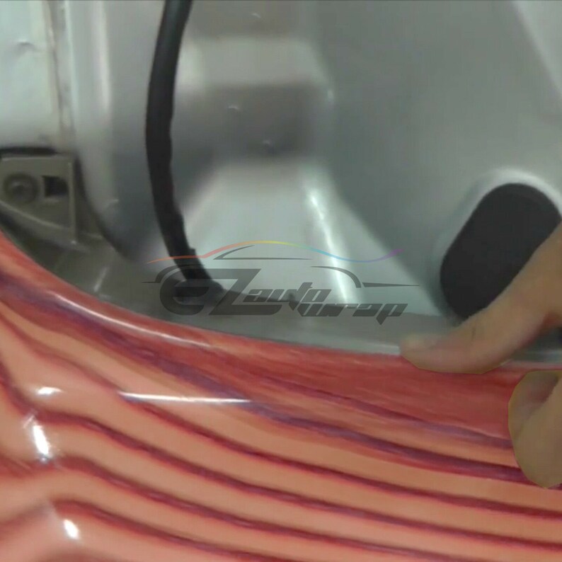 May include: Close-up of a wood grain vinyl wrap being applied to a car door. The wrap has a reddish-brown color with a wood grain pattern.