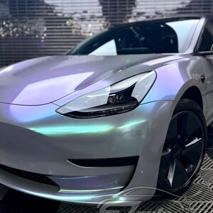 May include: A Tesla Model 3 sedan featuring a custom iridescent wrap that shifts between pearlescent white, soft purple, and mint green hues. The vehicle has a sleek front bumper, a sharp LED headlight assembly, and dark grey multi-spoke alloy wheels.