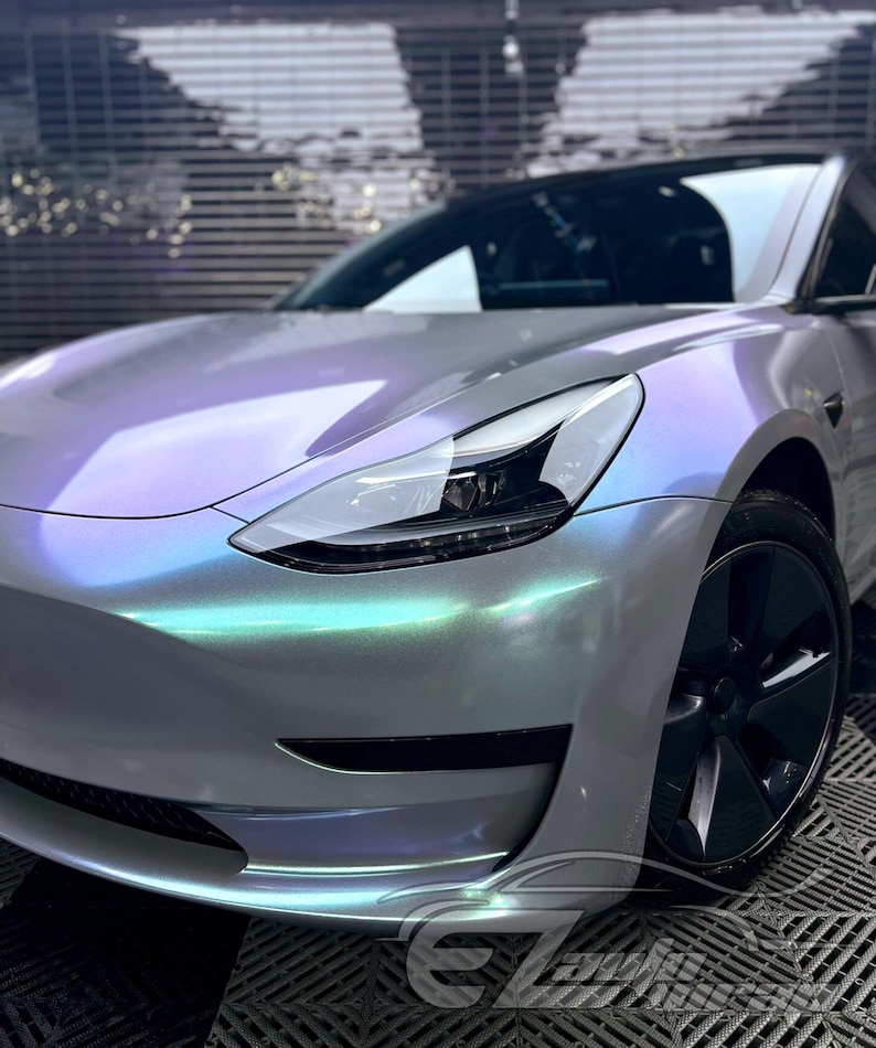May include: A Tesla Model 3 sedan featuring a custom iridescent wrap that shifts between pearlescent white, soft purple, and mint green hues. The vehicle has a sleek front bumper, a sharp LED headlight assembly, and dark grey multi-spoke alloy wheels.